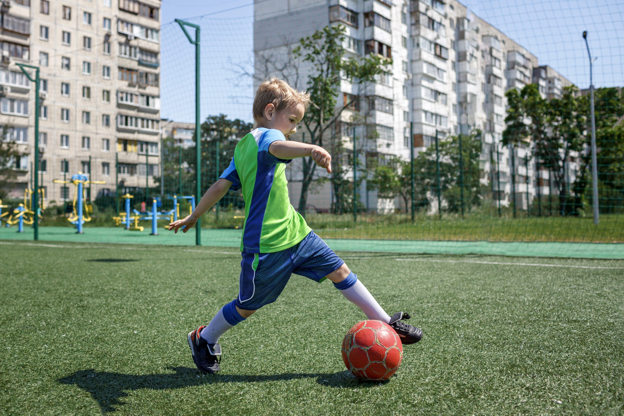 Little boy in blue and green form playing football on open field in the yard, a young soccer player